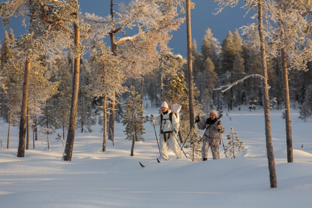 Kaksi lumipukuihin pukeutunutta metsästäjää hiihtää lumisessa maisemassa.